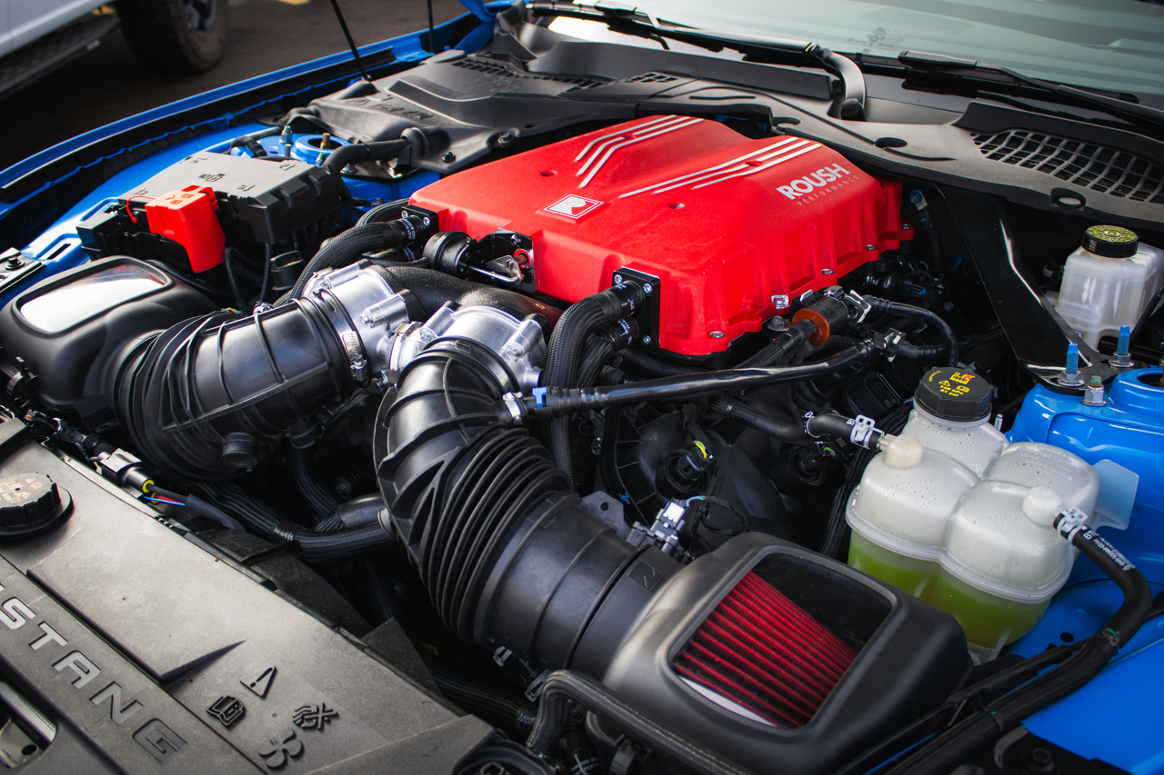 
                  
                    Close-up view of a car engine bay featuring the 2026 Roush Mustang Supercharger Kit installed on a Ford Mustang. This S650 Mustang supercharger delivers up to 810 horsepower, with various engine components and fluid reservoirs visible.
                  
                