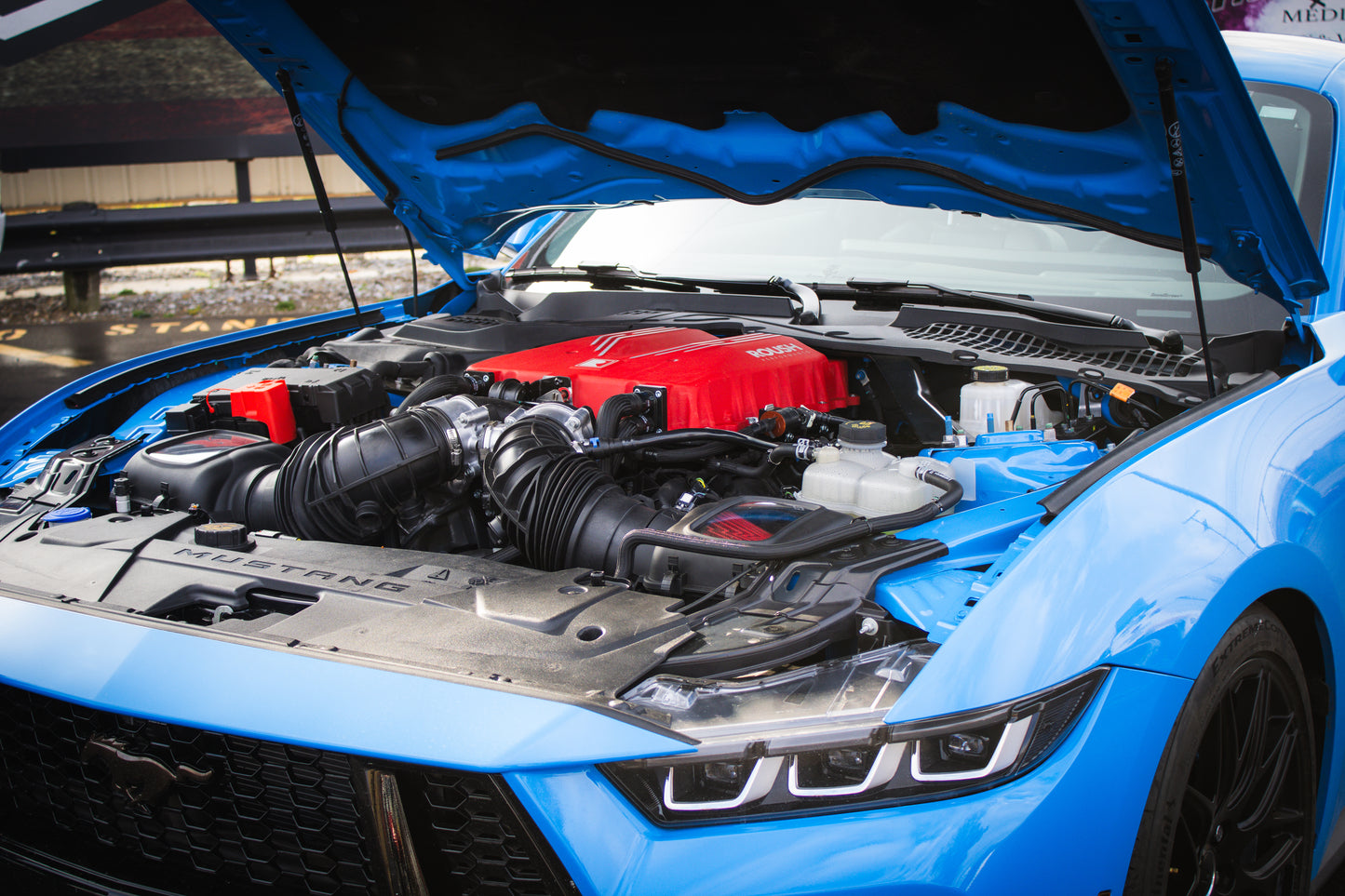 A blue Ford Mustang with its hood open, displaying a red engine cover and an 810 horsepower supercharger from the 2026 Roush Mustang Supercharger Kit.