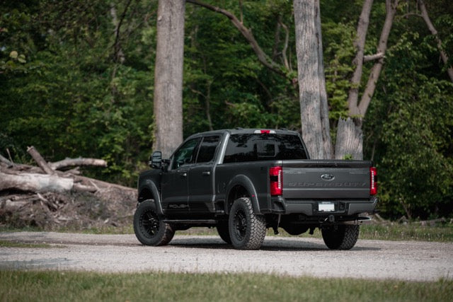 A black Ford F-250 pickup truck with off-road tires and performance suspension is parked on a gravel area in front of trees and greenery.