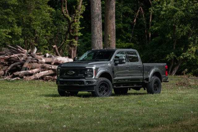 A gray 2023 Roush Super Duty Ford F-250 with performance suspension is parked on grass in front of a pile of logs, surrounded by trees and greenery.