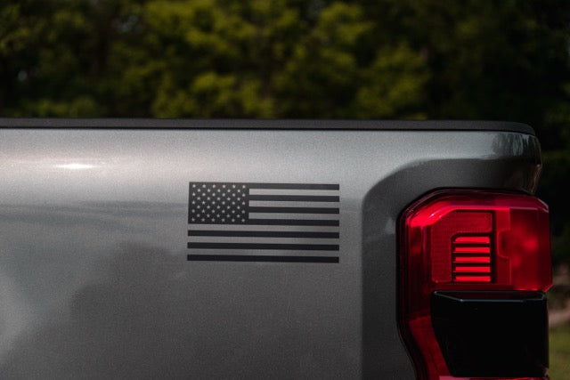
                  
                    Matte black American flag decal on the rear side panel of a gray Ford F-250 pickup, with part of the red taillight visible. Trees are blurred in the background, highlighting this 2023 Roush Super Duty’s bold look and performance suspension.
                  
                