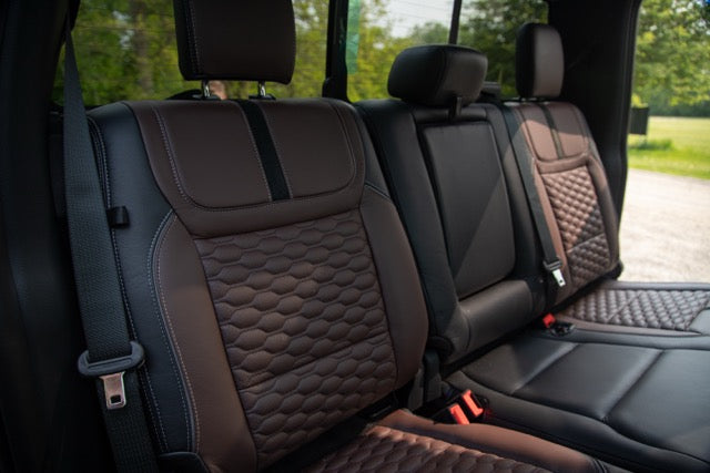 
                  
                    The image shows the back seats of a Ford F-250 with brown and black quilted leather upholstery and seatbelts, taken in daylight with trees visible outside the window—a glimpse inside a 2023 Roush Super Duty with premium comfort and style.
                  
                