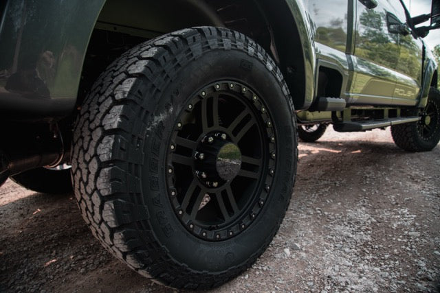 
                  
                    Close-up of a rugged off-road tire and black rim on a 2023 Roush Super Duty Ford F-250 with performance suspension, parked on a gravel surface.
                  
                