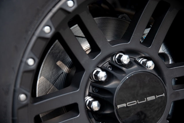 
                  
                    Close-up of a black alloy wheel with visible brake disc and lug nuts, featuring the "Roush" logo on the wheel center cap from the 2023 Roush Super Duty Ford F-250, highlighting its performance suspension upgrades.
                  
                