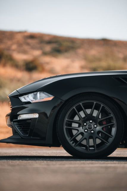 
                  
                    Close-up of the front left side of a black Roush Mustang parked on a road, with a blurred dry landscape in the background.
                  
                
