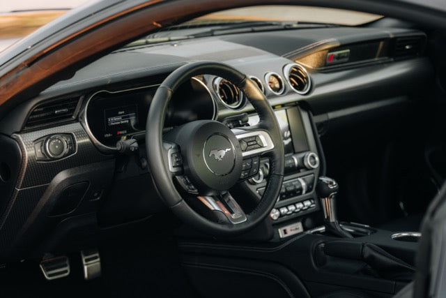 
                  
                    The image shows the interior of a modern Roush Mustang, featuring a black steering wheel with the Mustang logo, dashboard, and center console controls, highlighting its performance track car heritage.
                  
                
