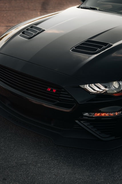 
                  
                    Close-up of the front end of a black Roush Mustang sports car, showing the grille, headlights, hood vents, and part of the bumper on a paved surface.
                  
                