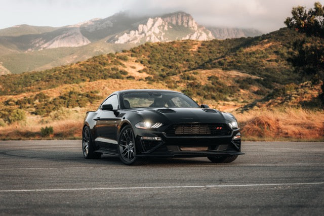 A black Roush Mustang is parked on an empty road with hills and mountains in the background under a partly cloudy sky.