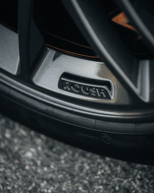 
                  
                    Close-up of a car wheel rim featuring the embossed "ROUSH" logo, with part of a tire and asphalt road visible in the background—perfectly capturing the spirit of a Roush Mustang performance track car.
                  
                