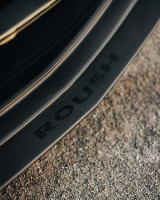 Close-up of a car part with the word "ROUSH" printed on it, positioned above textured pavement—highlighting the precision engineering of a Roush Mustang performance track car.