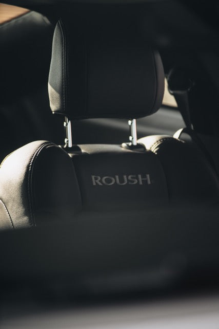
                  
                    Close-up of a black car seat headrest with the word "ROUSH" embroidered on the backrest, photographed inside a Roush Mustang performance track car.
                  
                