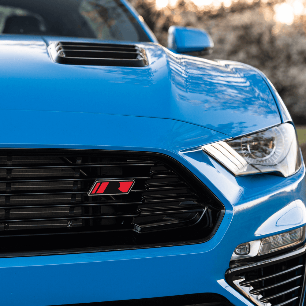 
                  
                    Close-up of the front of a blue sports car, showcasing the grille with a red "R" emblem, gloss black detailing, a prominent hood scoop with Mustang Hood Heat Extractors, and one of its headlights.
                  
                