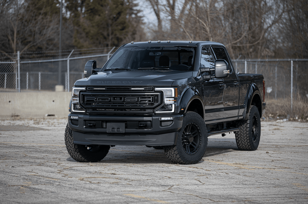 A black ROUSH F-150 pickup truck is parked on a paved surface, showcasing its high-flow front grille. In the background, a chain-link fence and leafless trees complete the rugged scene.
