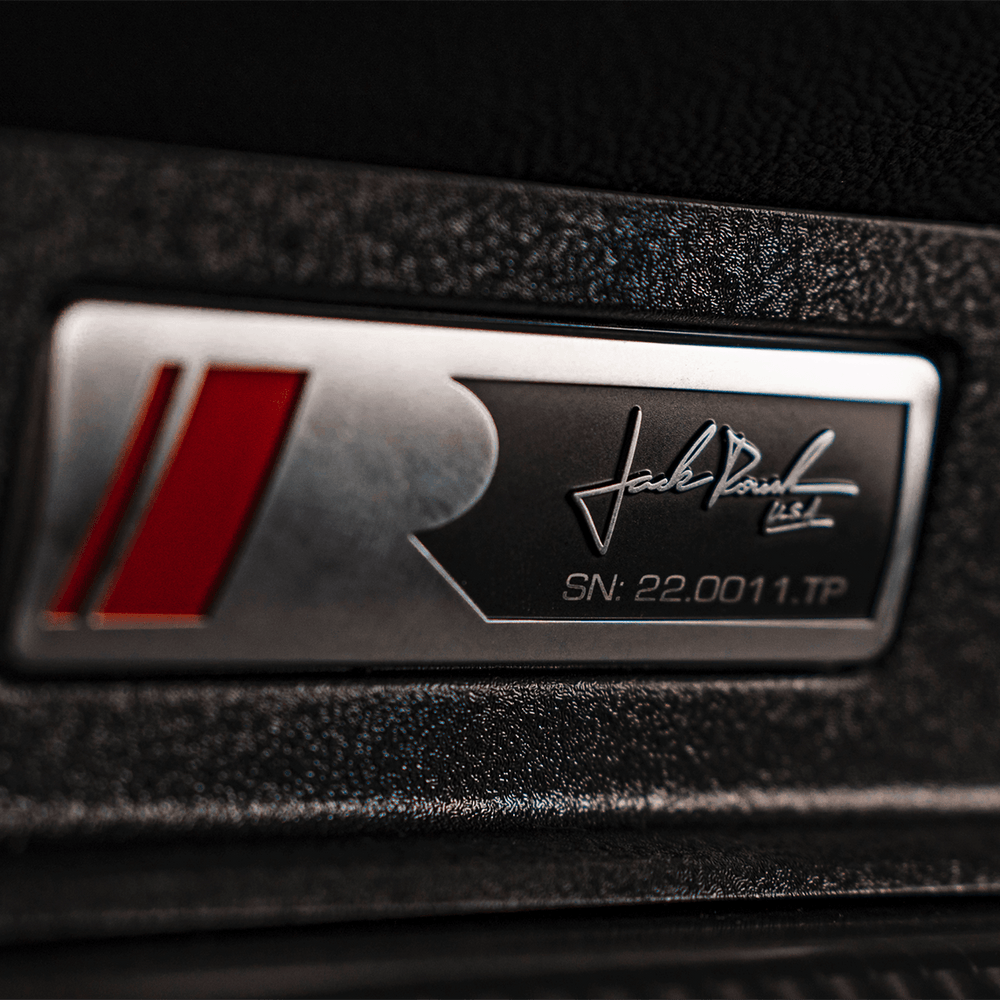 
                  
                    Close-up of a metallic badge with a red and silver "R" logo, a signature, and serial number "SN: 22.0011.TP" on a textured black surface, highlighting the elite craftsmanship associated with the performance-driven Roush Mustang.
                  
                