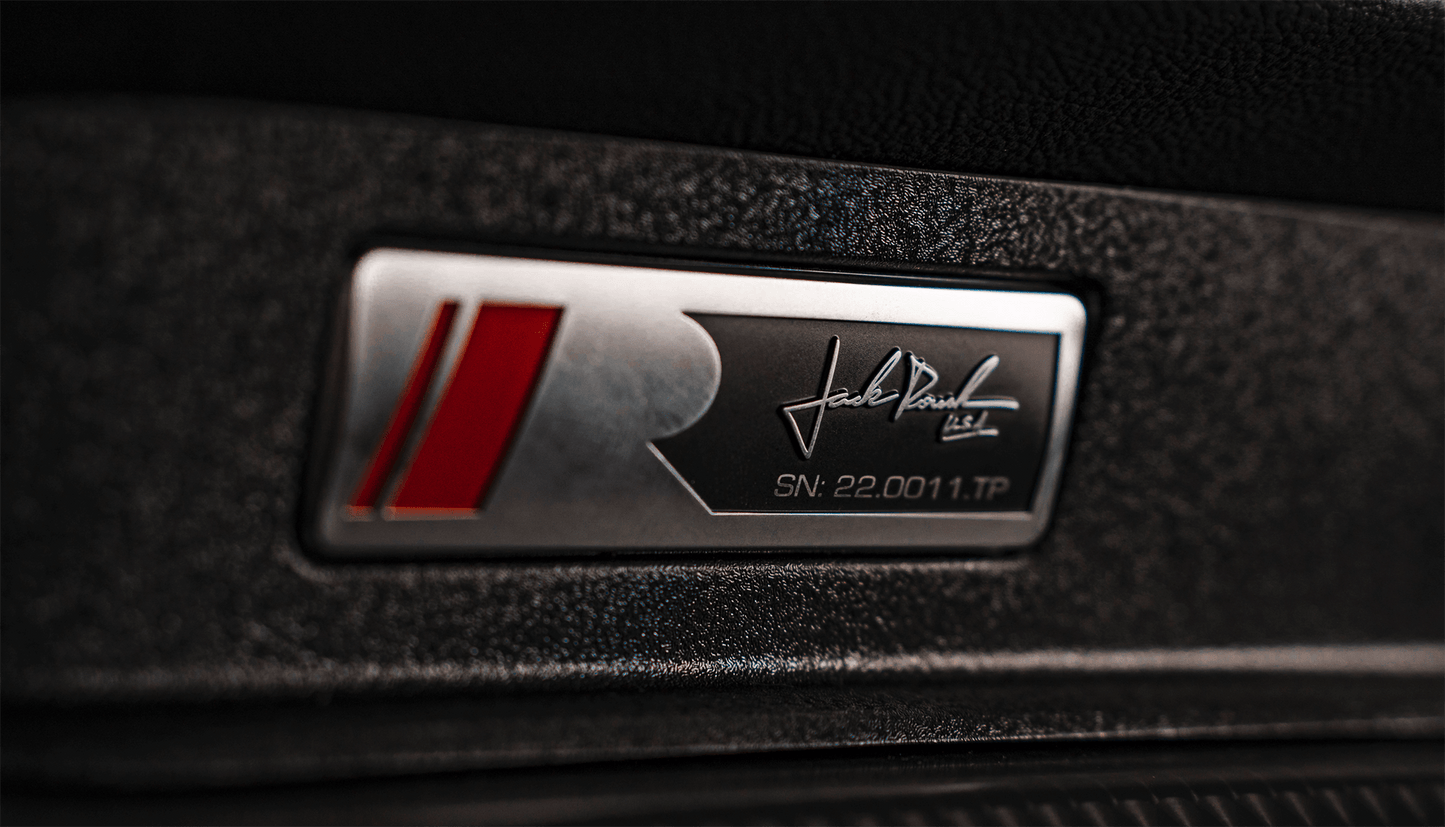 
                  
                    Close-up of a metallic badge with a red and silver "R" logo, a signature, and serial number "SN: 22.0011.TP" on a textured black surface, highlighting the elite craftsmanship associated with the performance-driven Roush Mustang.
                  
                