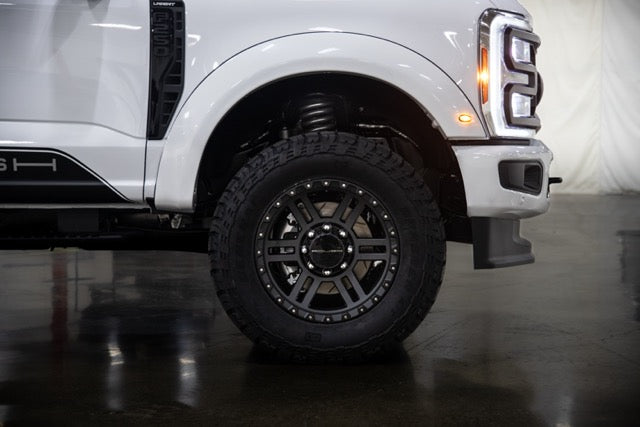 
                  
                    The front left wheel and fender of the white pickup truck, featuring rugged tires and a performance suspension, are parked indoors on a reflective surface.
                  
                