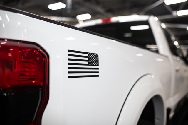 
                  
                    Close-up of a white pickup truck featuring a striking black and white American flag decal near the rear, parked indoors. This Roush Super Duty boasts a powerful turbodiesel V8 engine and performance suspension for an unparalleled driving experience.
                  
                