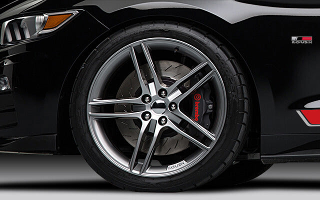 
                  
                    Close-up of a car wheel featuring a silver alloy rim, Brembo brake caliper, and a Roush badge, showcasing the sleek design of the ROUSH Mustang's Stage 1 package against its black car body.
                  
                